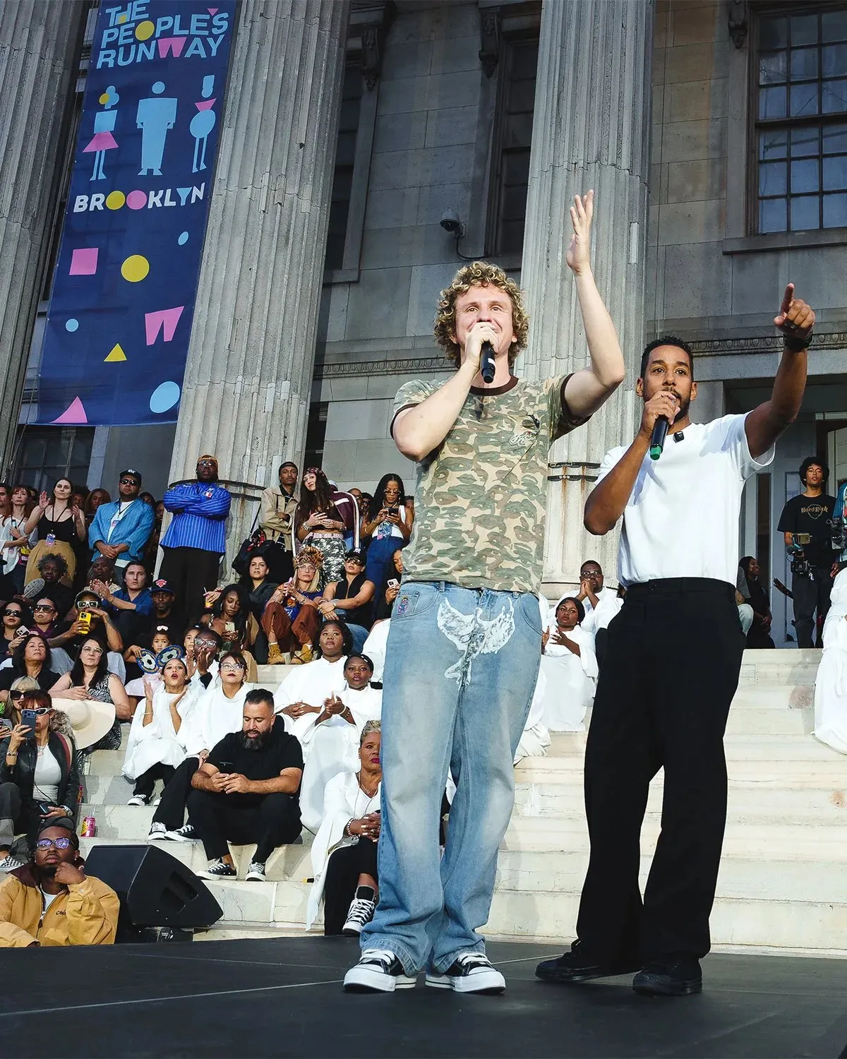 Colm Dillane and another presenter address the crowd from the steps of Brooklyn Borough Hall beneath a People’s Runway banner