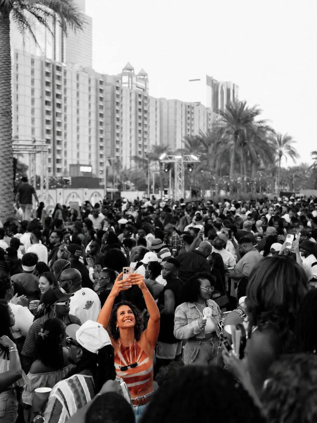 A festival-goer in an orange top raising a phone above a monochrome crowd at The Cookout