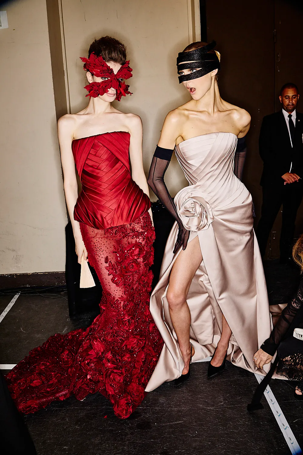 Two models backstage in sculptural red and ivory gowns, wearing feathered and black masks as one turns toward the other