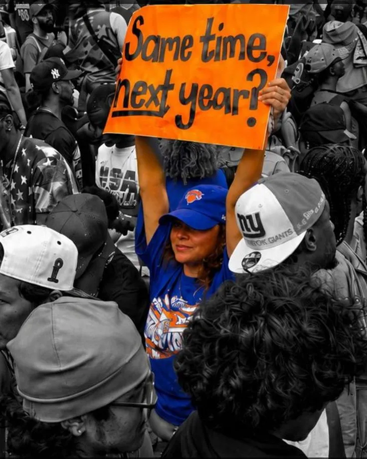 A fan in Knicks blue holding an orange "Same time, next year?" sign above a monochrome celebrating crowd