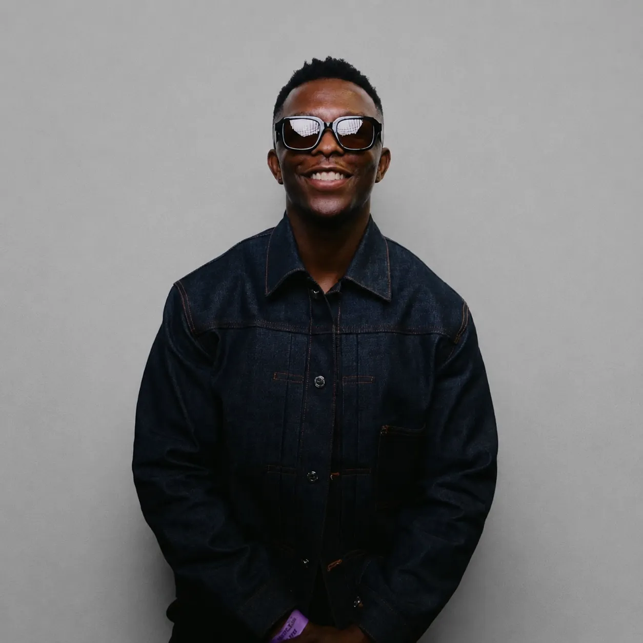 Tolu Farinto smiling in sunglasses and a dark denim jacket against a plain grey wall