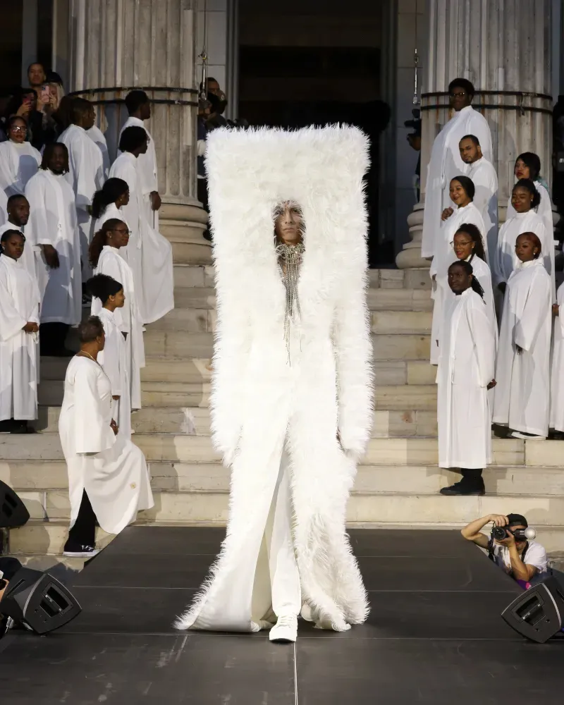 A model in a floor-length white sculptural coat and white Converse sneakers walks the runway on the steps of Brooklyn Borough Hall between rows of performers dressed in white