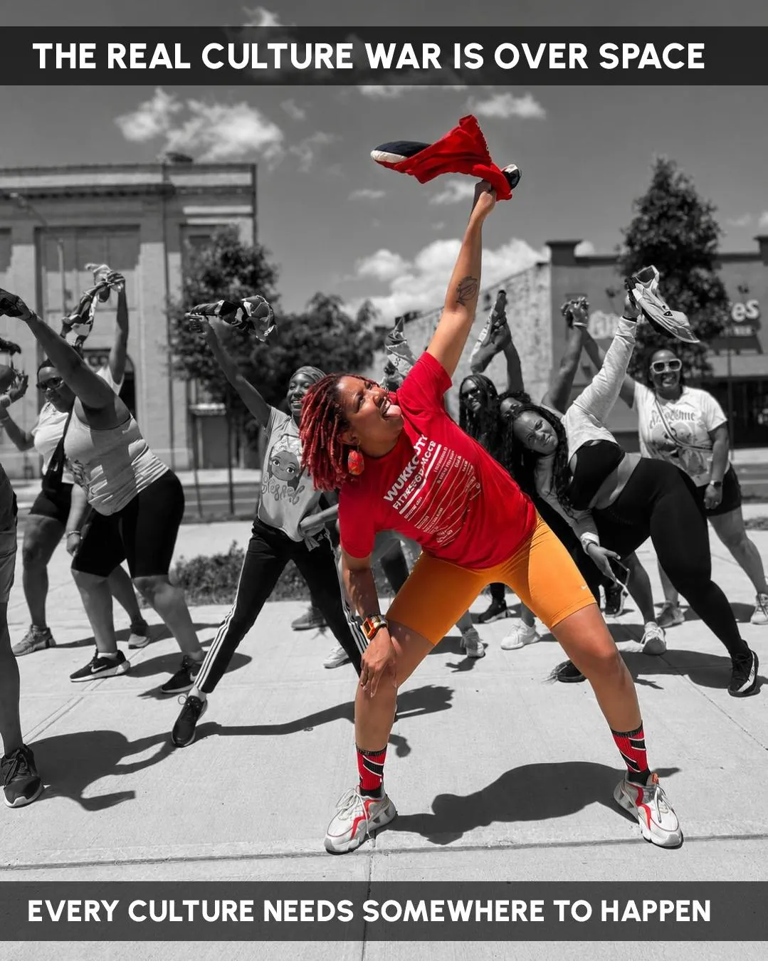 A dance fitness class on a Brooklyn sidewalk, the leader's red shirt and orange shorts in colour against a monochrome street