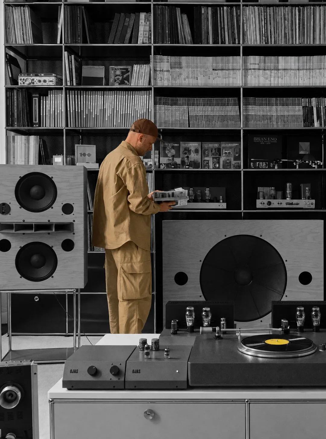 A man in tan workwear selecting music inside a monochrome OJAS listening room, with a yellow record in the foreground