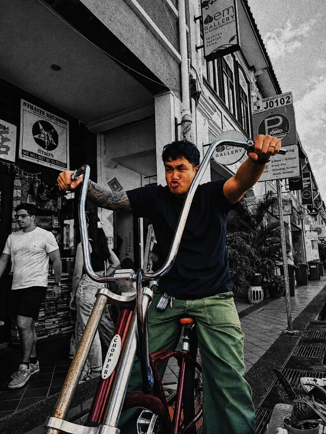 A tattooed man leans over a tall-handlebar bicycle outside The Panic Room, his skin, green trousers and red bike picked out in colour